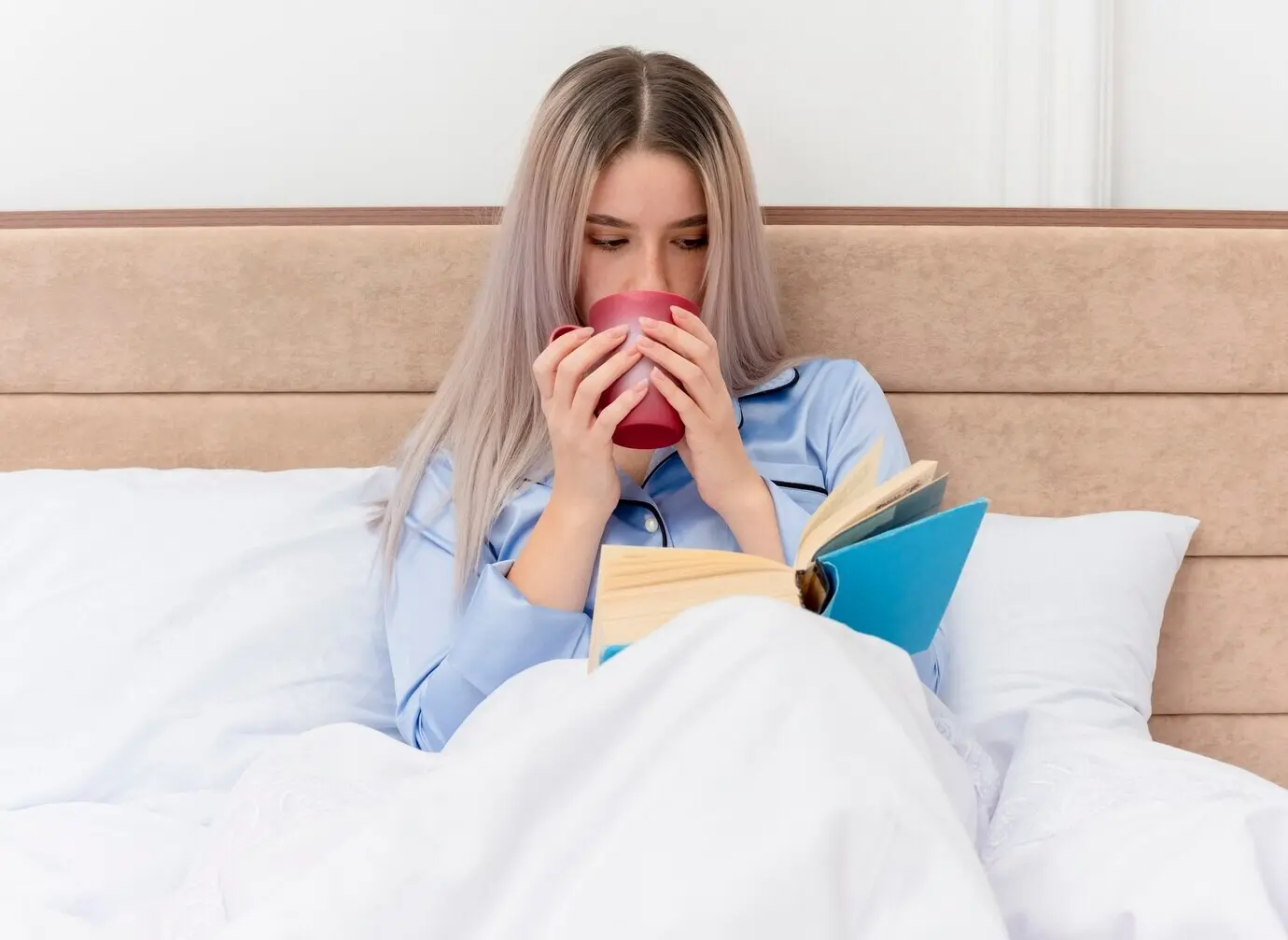 Eine junge, schöne Frau im blauen Pyjama sitzt im Schlafzimmer auf dem Bett mit einer Tasse Kaffee und liest ein Buch vor hellem Hintergrund.