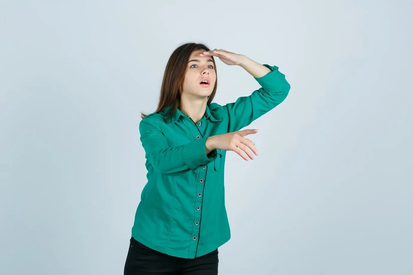 Junges Mädchen mit der Hand über dem Kopf blickt konzentriert in die Ferne und zeigt von sich weg, in grüner Bluse und schwarzer Hose, Vorderansicht.